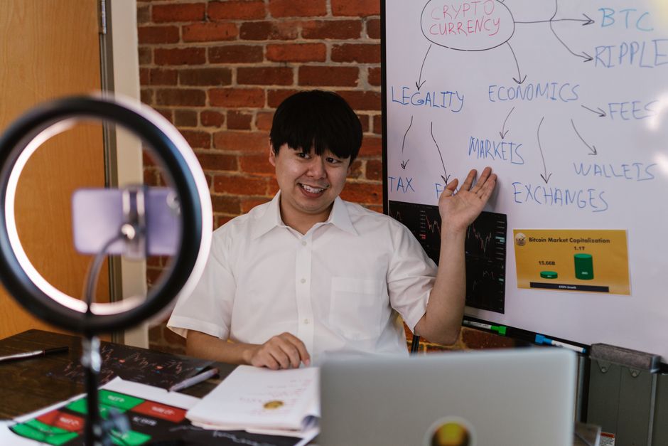A man in a white shirt sits at a desk in front of a brick wall, gesturing towards a whiteboard filled with diagrams and notes related to economics and cryptocurrency. The desk has various papers, a la