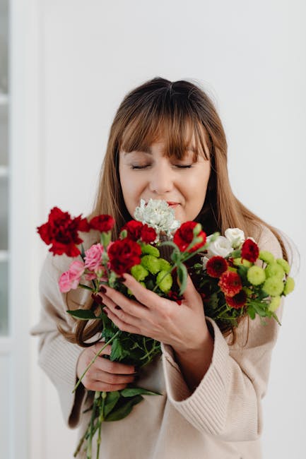 A woman with light brown hair and bangs, dressed in a beige sweater, is holding a vibrant bouquet of fresh flowers close to her face with her eyes closed. The bouquet features a variety of blooms, inc