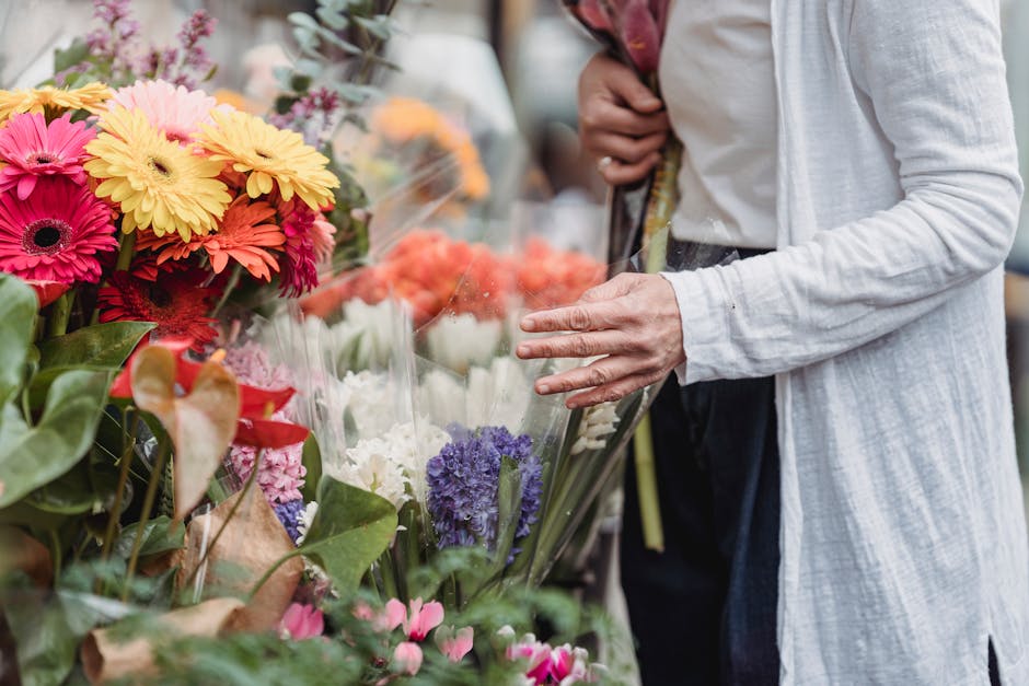 A person wearing a white long-sleeved shirt and dark trousers selecting fresh flowers at a flower market. The bouquet features vibrant gerbera daisies in shades of pink, orange, and yellow, along with