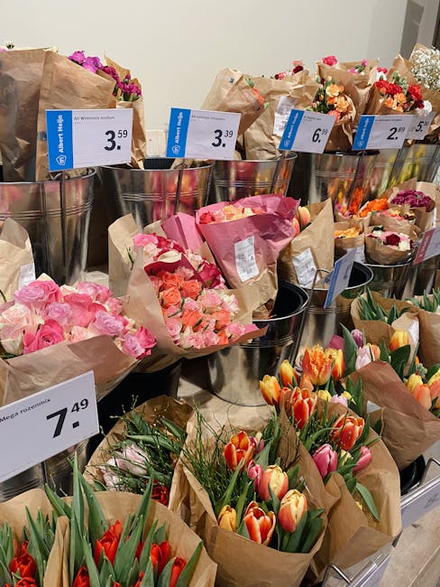 A variety of bouquets arranged in metal buckets and wrapped in brown craft paper, displayed on a retail shelf. The bouquets include pink roses, orange and pink tulips, and multicolored carnations, all