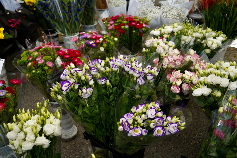 A vibrant display of fresh flowers arranged in bouquets and floral bundles on a flower shop table. The collection includes purple and white lisianthus, pink and white roses, red and pink chrysanthemum