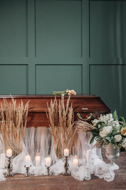A floral arrangement featuring white roses, peonies, and greenery in a white vase, placed on a wooden surface in front of a green paneled wall. Surrounding the vase are tall, dried pampas grasses and 