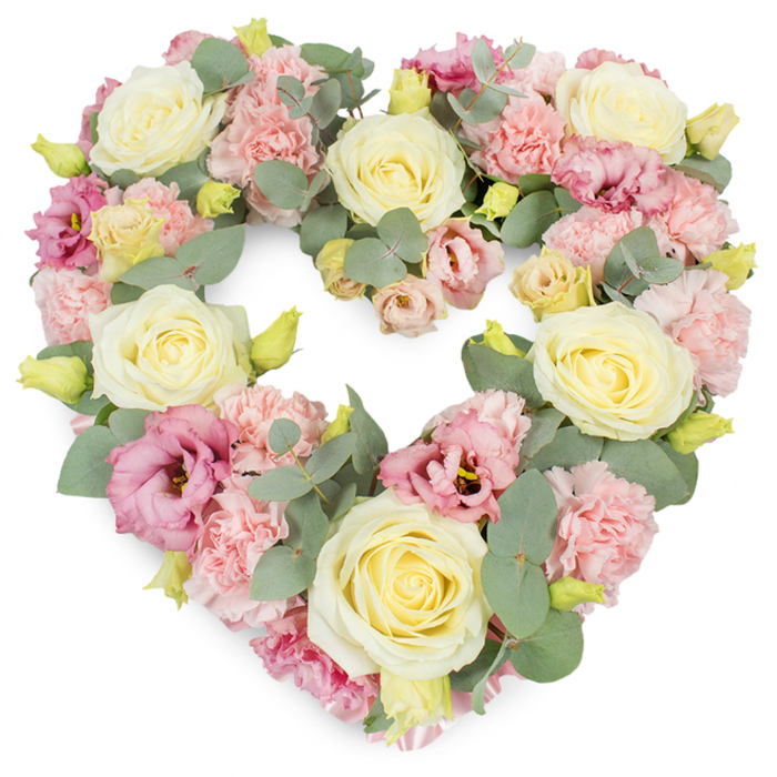 A full, symmetrical heart-shaped wreath rests against a bright white background, its pastel blooms combining to create an atmosphere of peace and loving remembrance, perfectly suited to the close-knit community of Biggin Hill. The heart itself is clearly defined, with the floral outline smooth and unbroken, suggesting unity and the continuity of cherished memories. Large, creamy ivory and soft yellow roses are positioned around the form, their spiralling centres and outer petals opening just enough to show depth without appearing overly blowsy. These roses act as anchor points in the design, drawing attention and symbolising affection and respect. Filling the spaces between them are generous groupings of light blush-pink carnations; their multi-layered, ruffled petals lend a soft, tactile quality, like folded silk or lace. Interspersed among the roses and carnations are deeper pink to mauve lisianthus blooms, some with slightly darker centres that add visual interest and an elegant, almost romantic touch. Woven throughout the design, sprigs of silvery eucalyptus foliage introduce cool, muted greens; their rounded leaves overlap and fan out, framing the flowers and giving the wreath a natural, restful feel that echoes the nearby green spaces and airfield horizons. Tiny yellow-green buds appear at intervals, close to the larger blossoms, adding a sense of newness and life. The lighting used in the photograph is gentle yet clear, picking up the subtle variations in tone and texture while keeping the overall effect soft. This heart-shaped tribute would be chosen by families in Biggin Hill seeking to convey sympathy, comfort, and unending love at a funeral, graveside service, or memorial occasion.